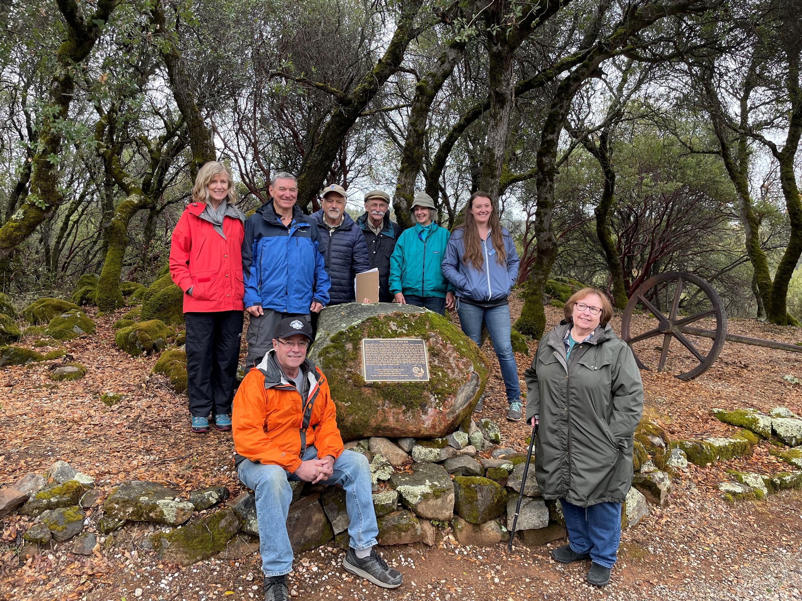 A small group of people gather around a plaque embedded in a small granite boulder covered with moss