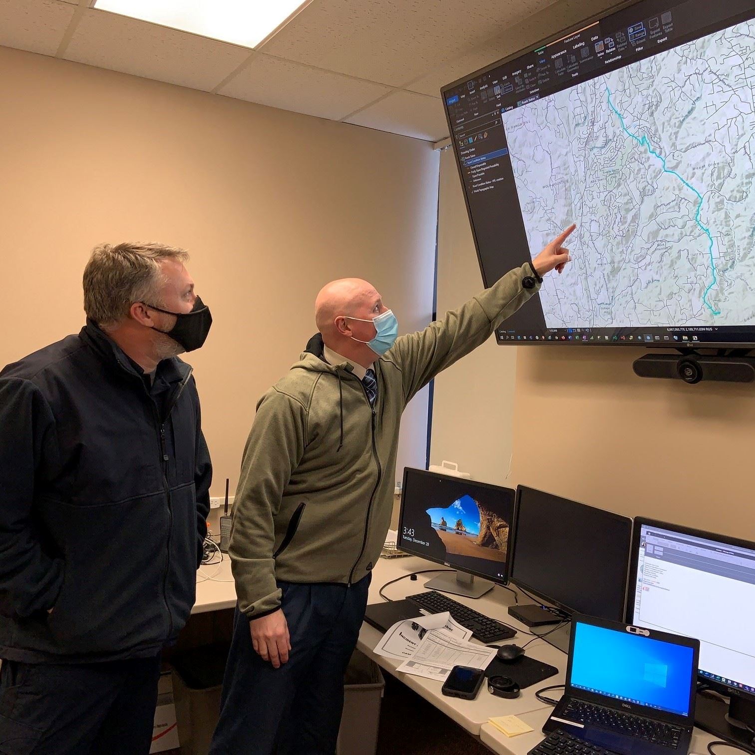 Two men look at a large map on a wall-mounted TV screen. One of the men points at the map.