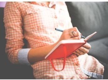mid section of woman with red notebook in lap, checkered orange shirt 