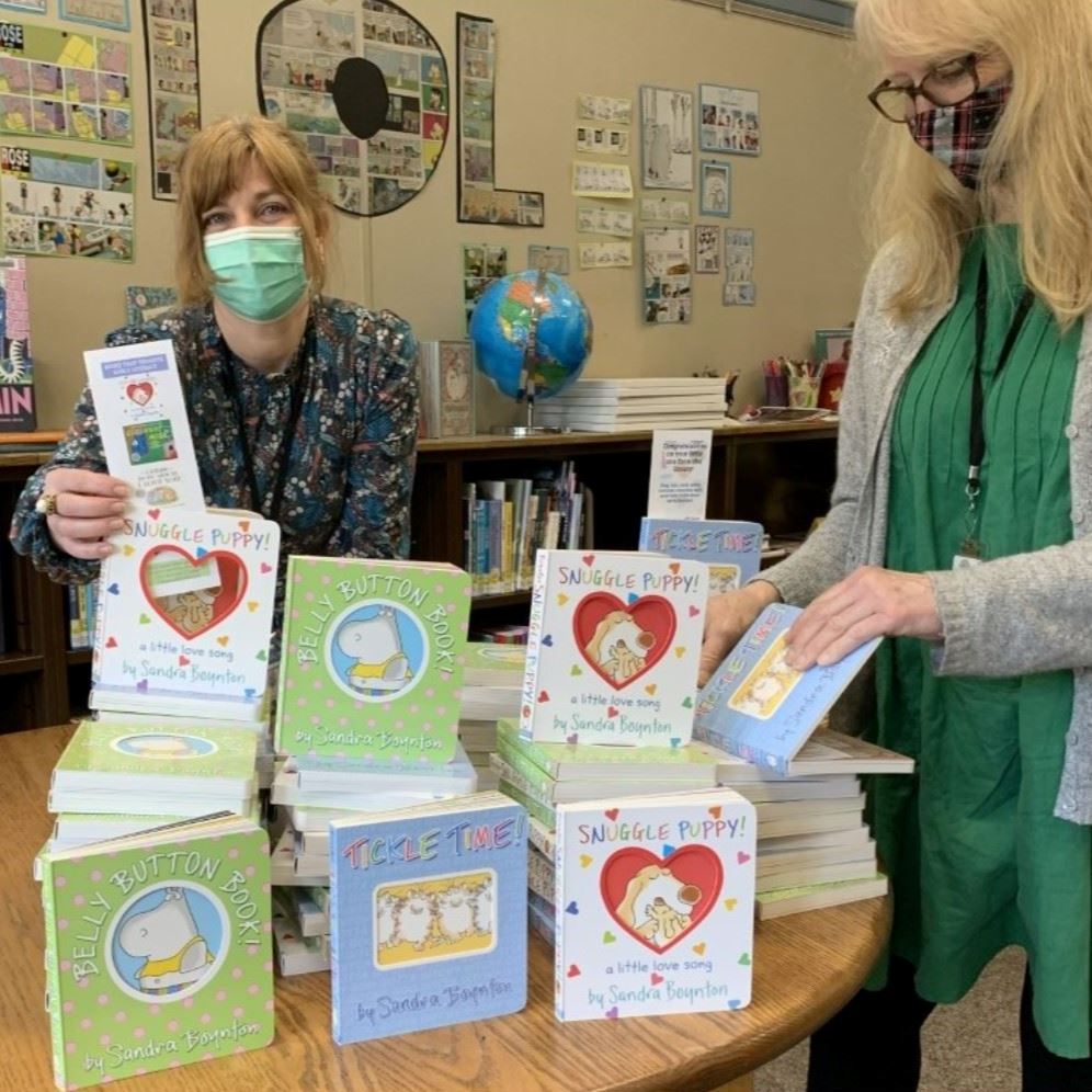 Librarians Jill and Teri show off a stack of pastel board books for newborns