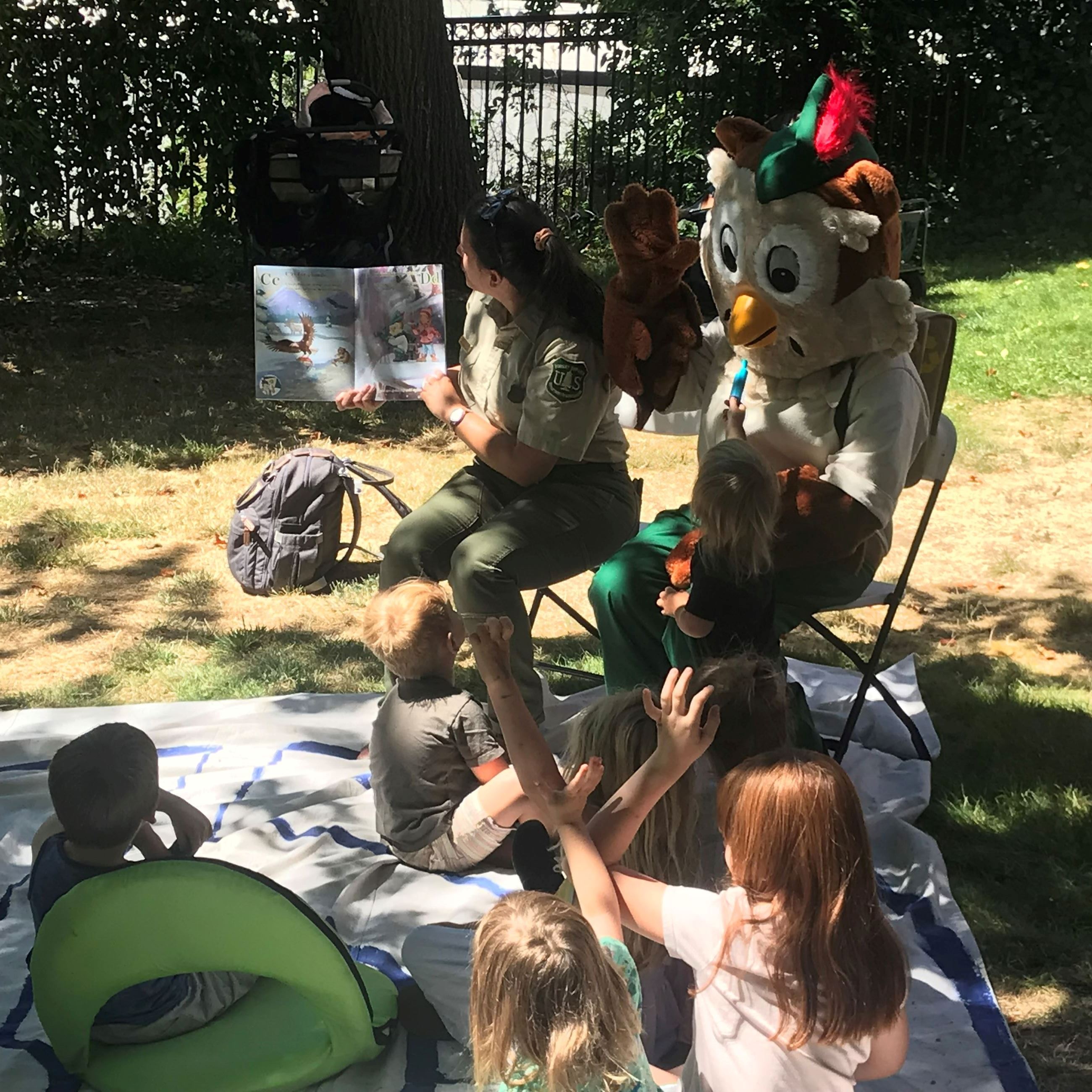 Woodsy the Owl reads a picture book to a group of children in a sunny backyard