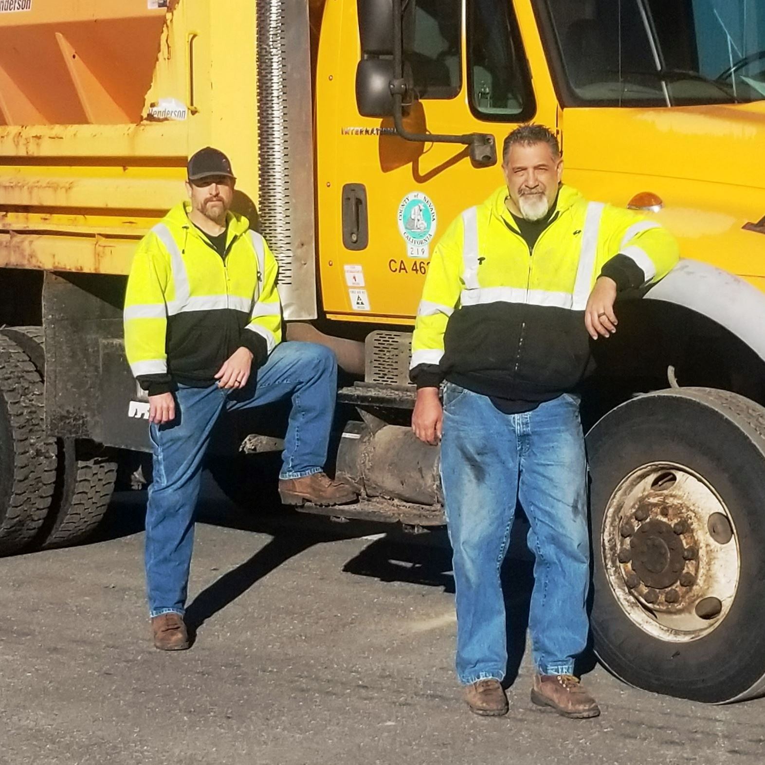 Two people dressed in bright yellow jackets lean against a bright yellow dump truck