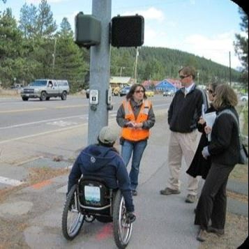Pedestrians, three on foot and one using a wheelchair, wait to cross at an intersection