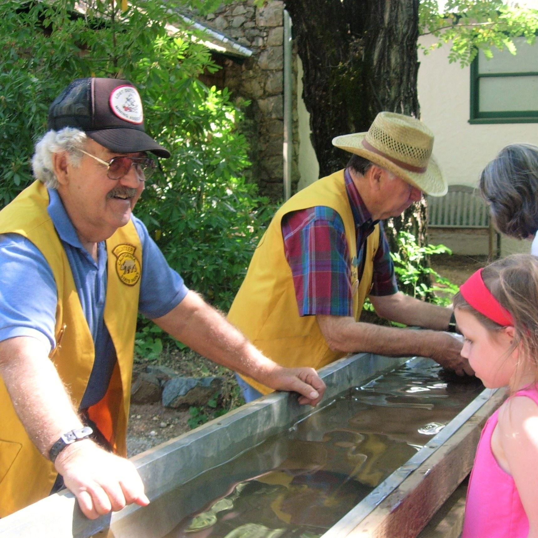 A person wearing a gold vest leans over a trough of water to speak to a child in a pink headband