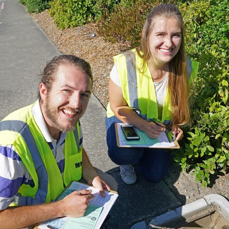 Two young people wearing bright yellow vests and holding clipboards crouch over a water main
