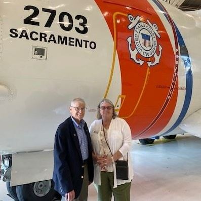 Two people stand in front of a large white and red Coast Guard airplane in a hangar