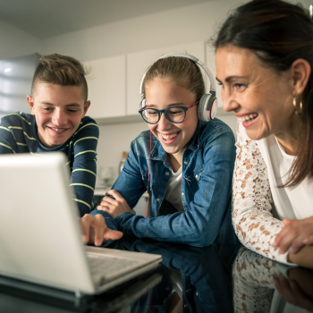 A teen wearing headphones looks types on a laptop as their sibling and parent watch smiling