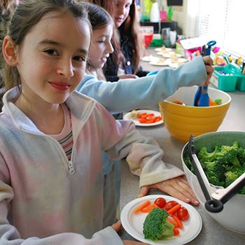 white child with brown hair smiling with a bowl of vegetables in her hand