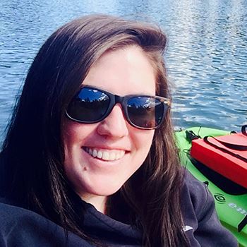 smiling white woman with brown hair and sunglasses on with a lake and boat in the background