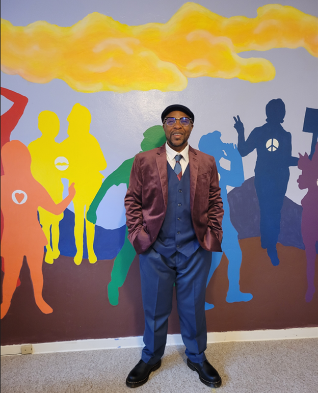 Callum Hancock wearing blue slacks and vest  with a tie and brown velvet sport coat and a beret